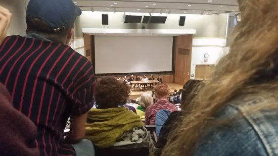 A back row view of a full audience of people sitting in dye lecture hall. On the stage is a table with 6 professor sitting