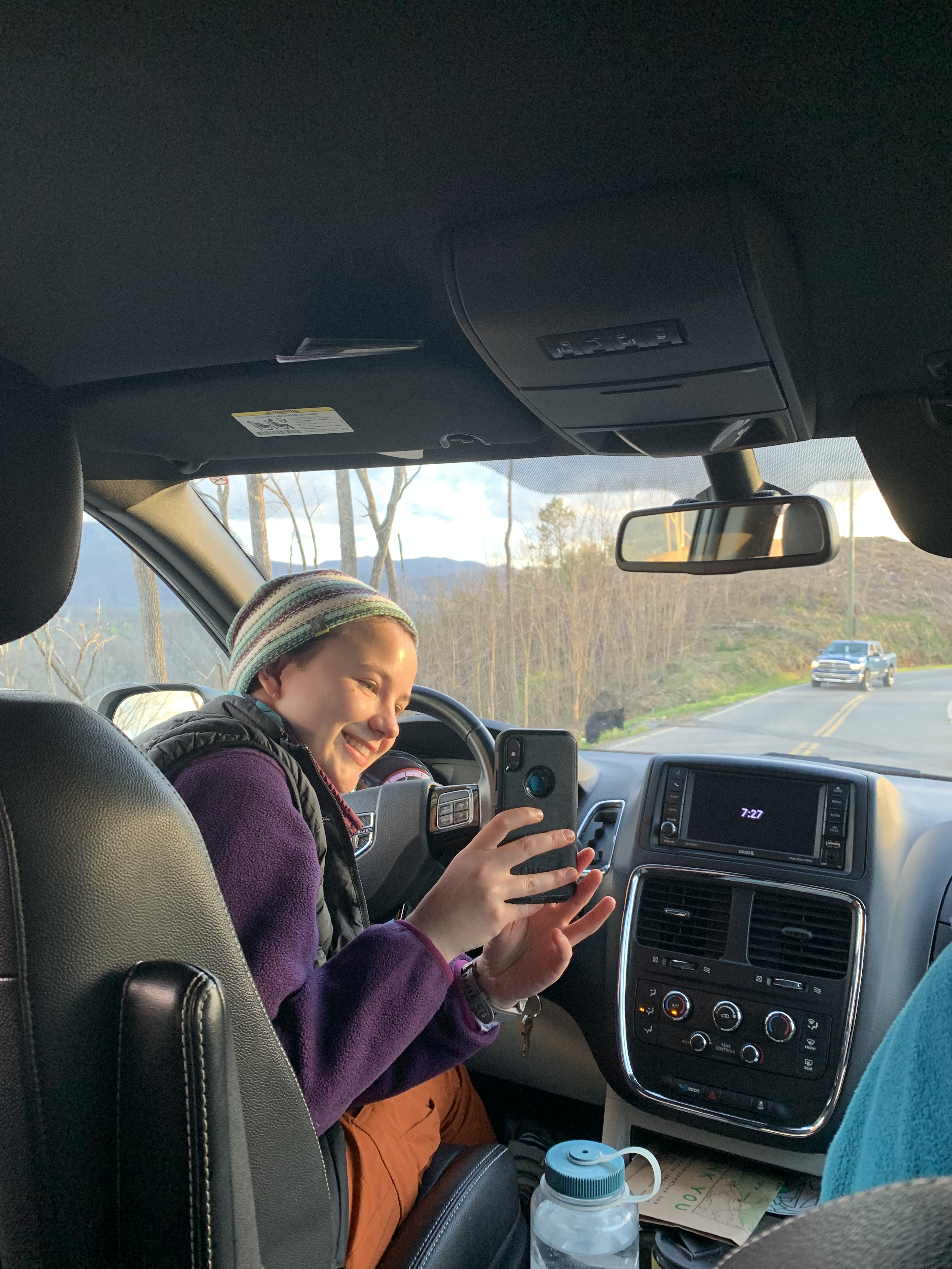 a person sits in the driver's seat of a car and holds out their phone to take a selfie of the road in front of them. there is a small black blob on the side of the road (it is a bear)