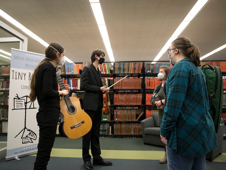 Two students holding a violin and guitar speak with fellow students in a library.