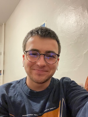Sionainn, a white transmasculine person with short dark hair, is sitting in their room. They are wearing dark glasses, headphones, and a blue shirt. They are grinning into the camera for a #selfie moment. 