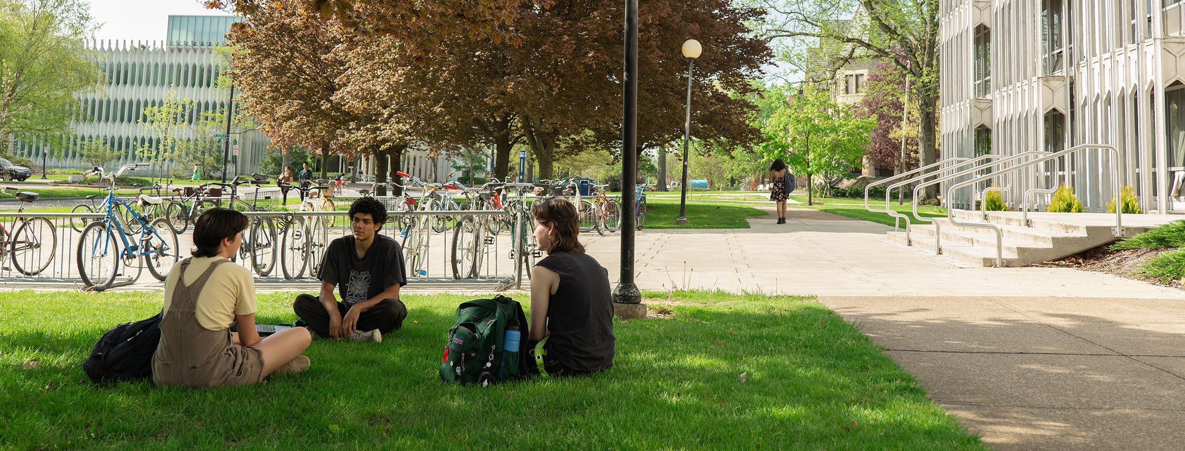 Seated in a circle in the grass, several students work on laptop computers. All are masked.