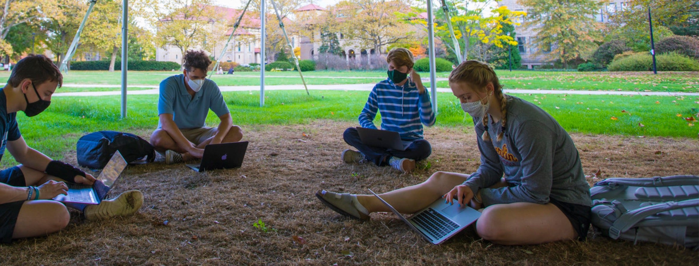 Seated in a circle in the grass, several students work on laptop computers. All are masked.