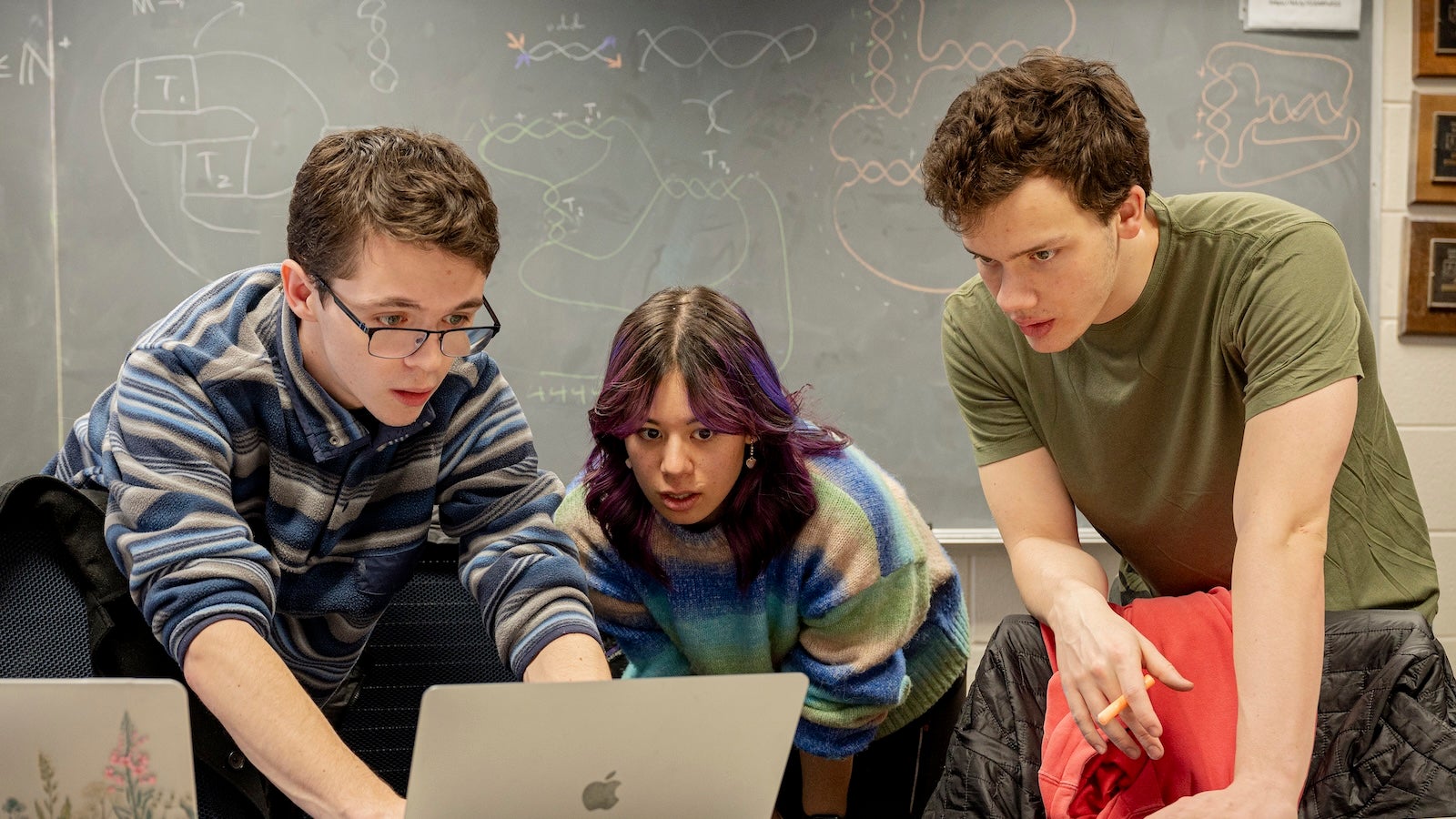 Three students lean over a table, looking at a laptop screen.