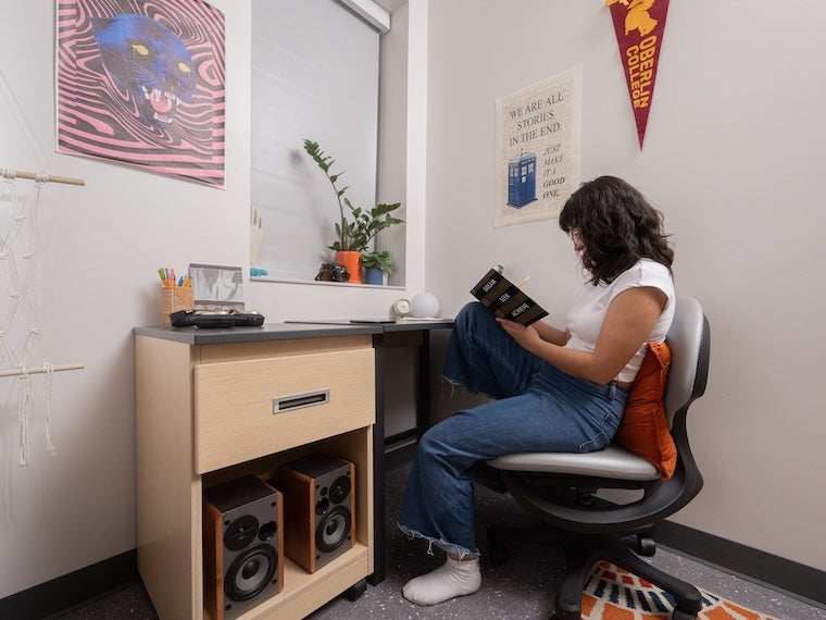Student sitting at a desk in a dorm room, reading a book.