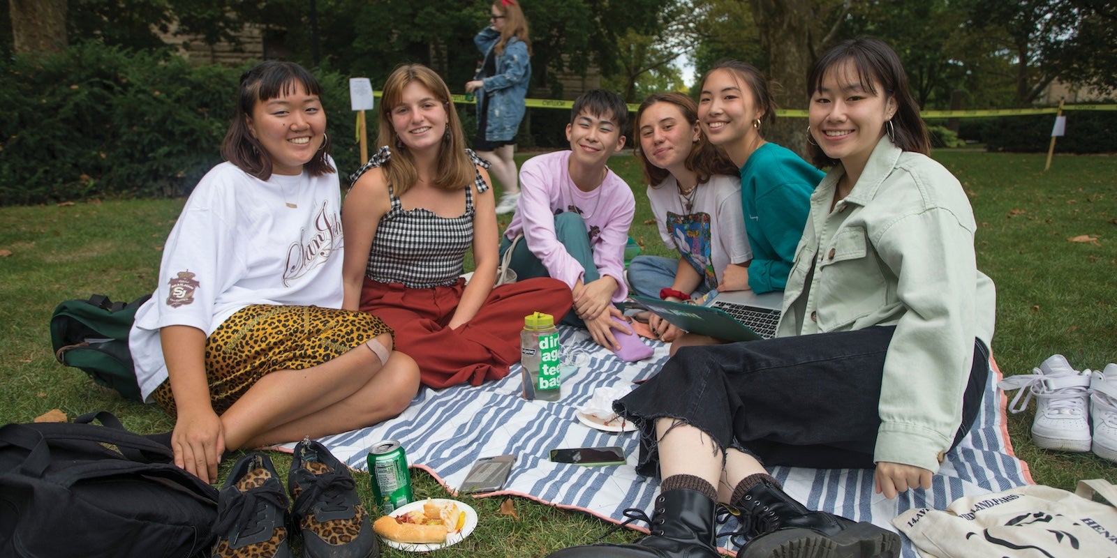 A group of friends sitting on a blanket outdoors, smiling and enjoying food and drinks together.
