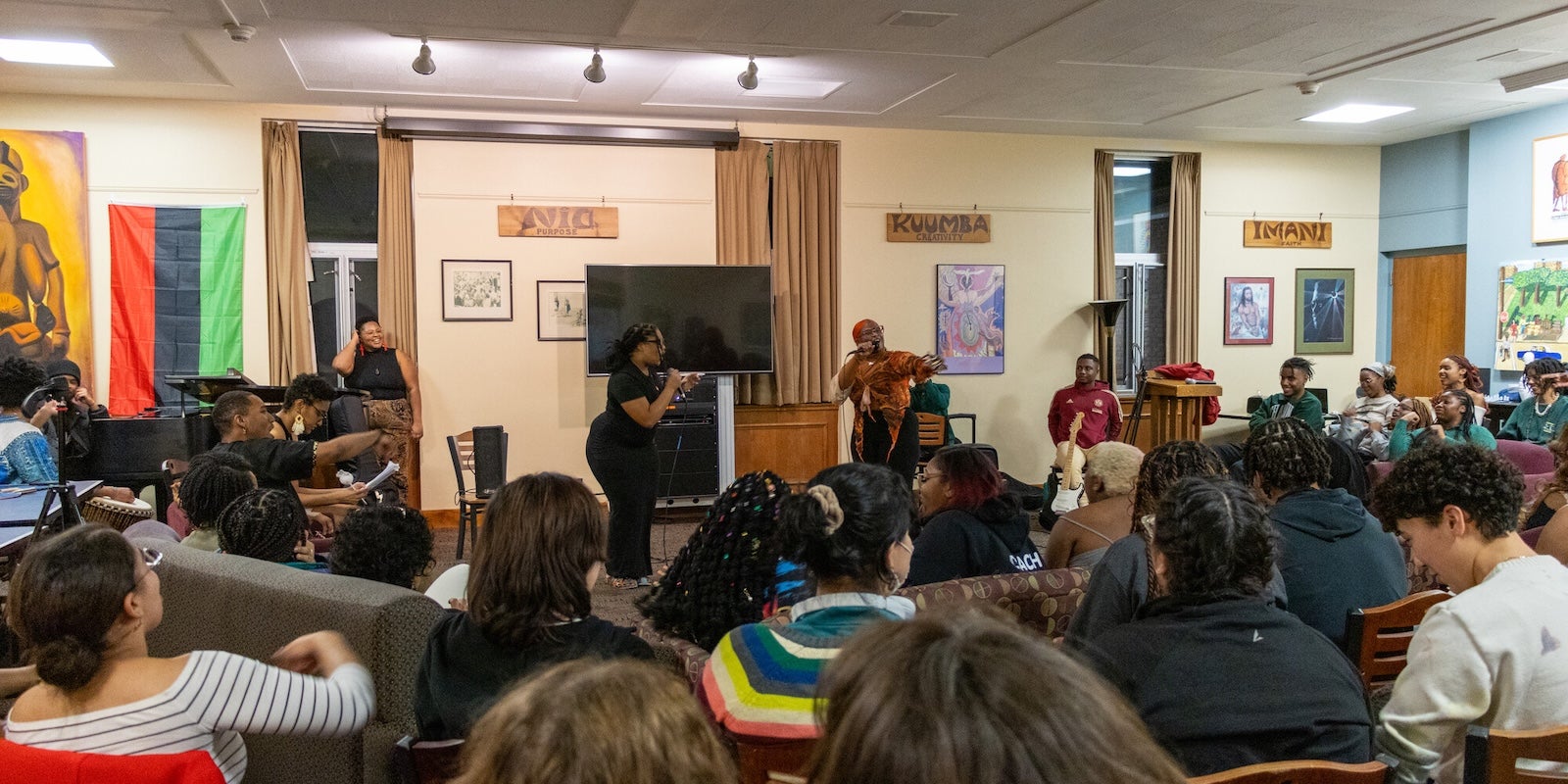 Students gathered in a lounge for a lively performance or discussion, with speakers engaging the audience at the front of the room.