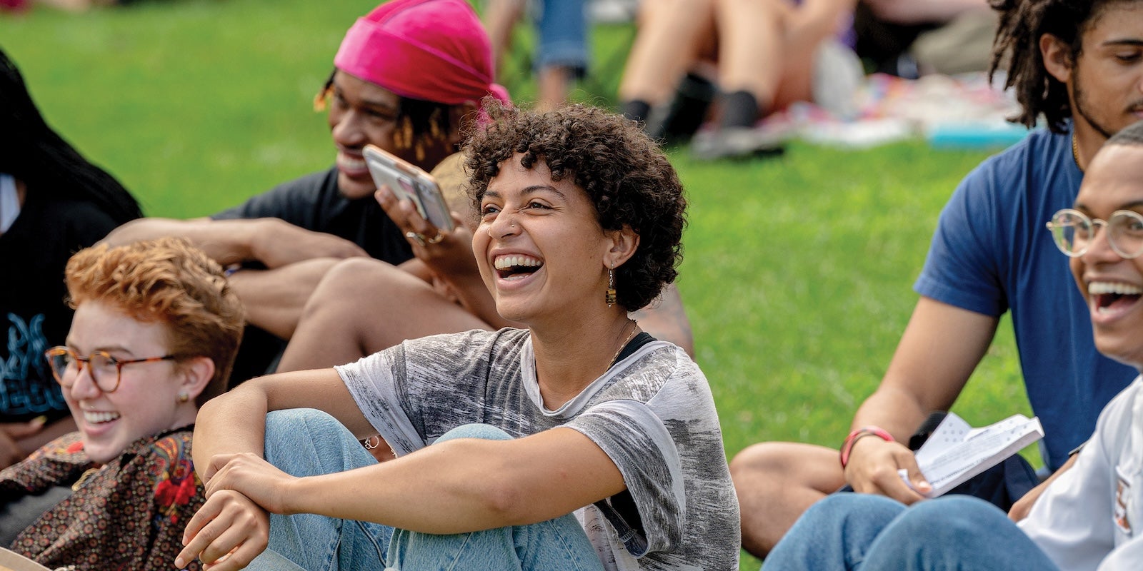 A group of students sitting on grass outdoors, laughing and enjoying a casual gathering.
