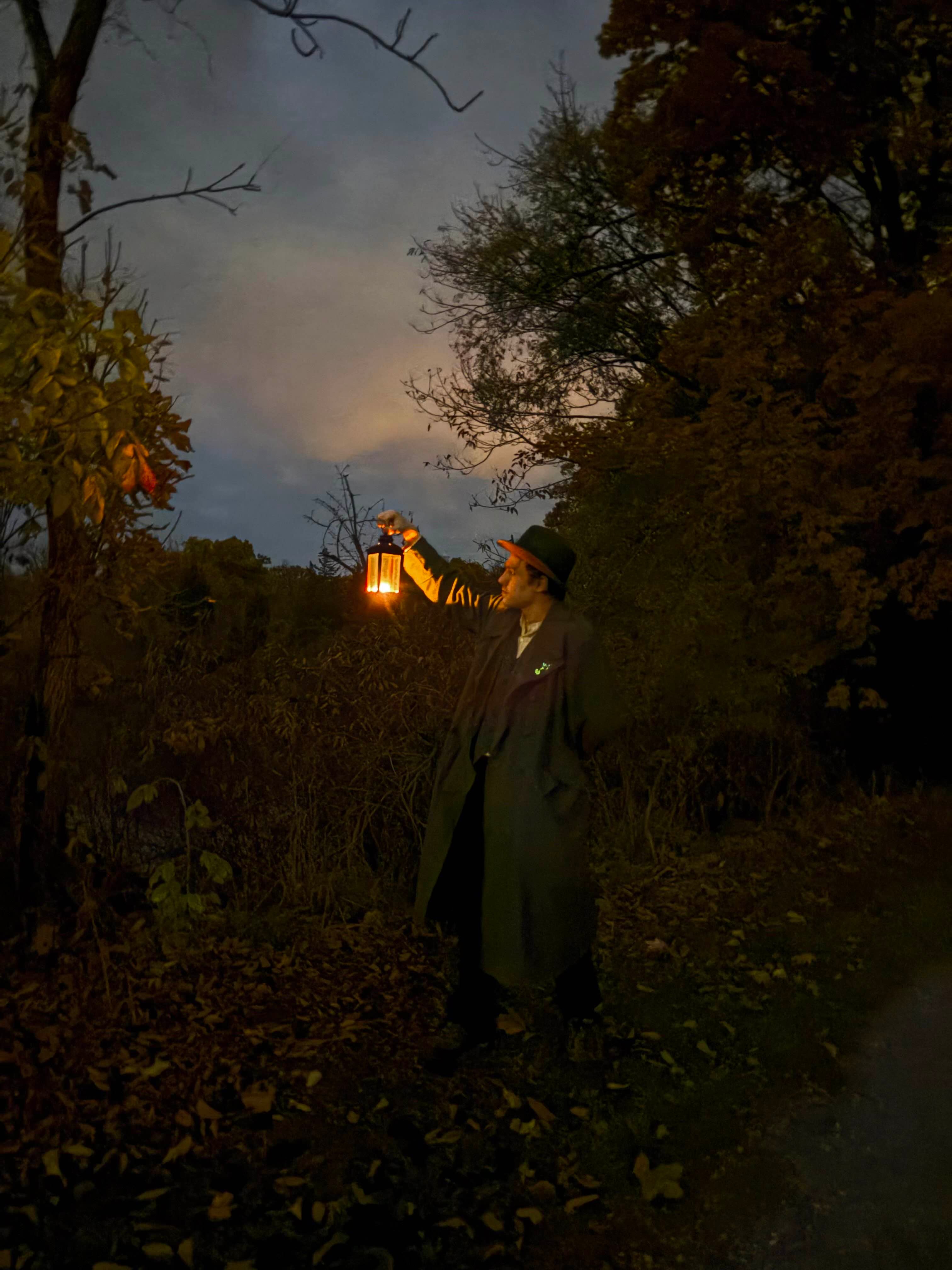 A figure in a coat stands in the Oberlin Arboretum at night, holding a lantern.