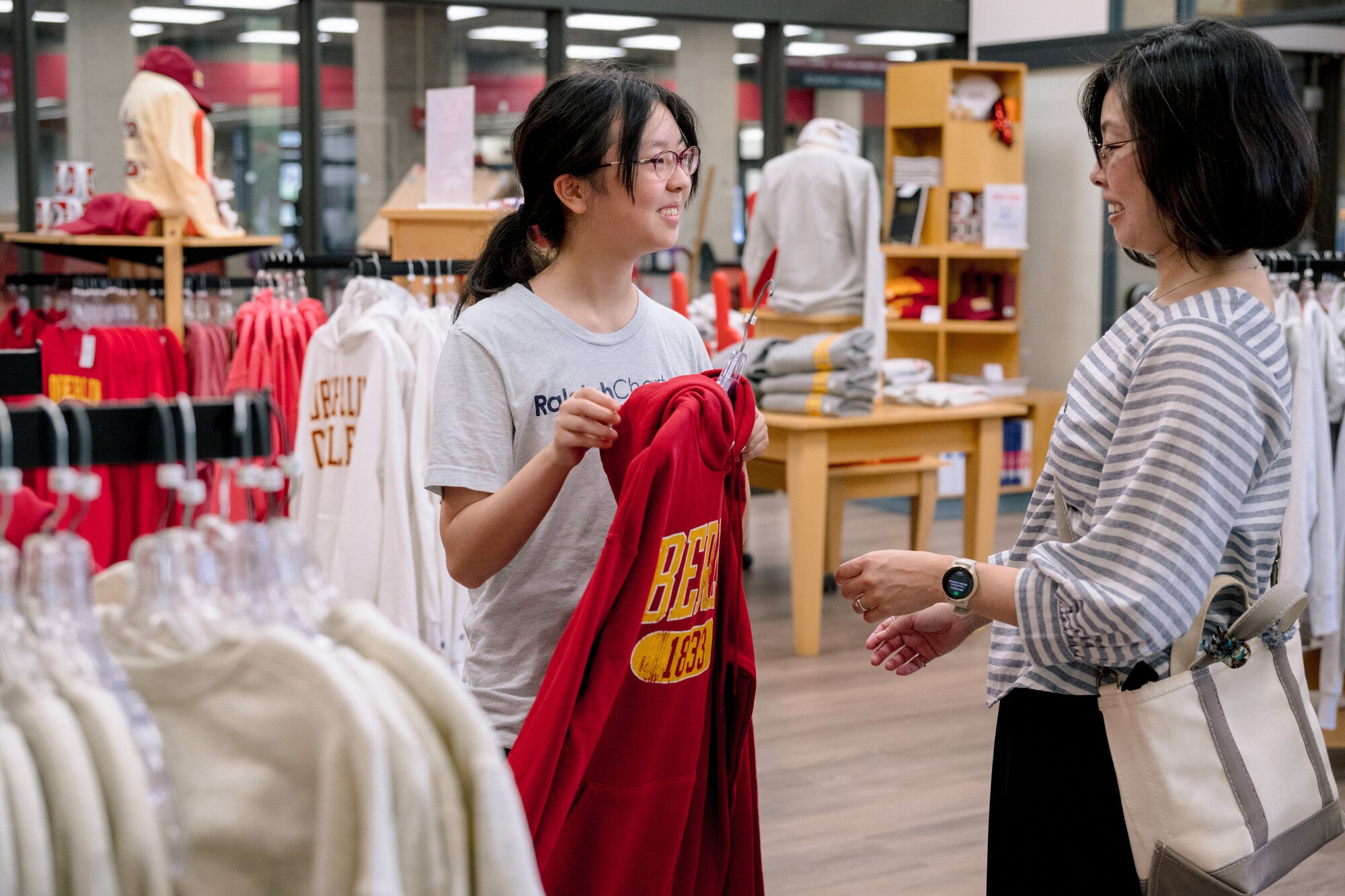 A parent and a student inside a campus store, the student holding up a red sweat shirt while talking.