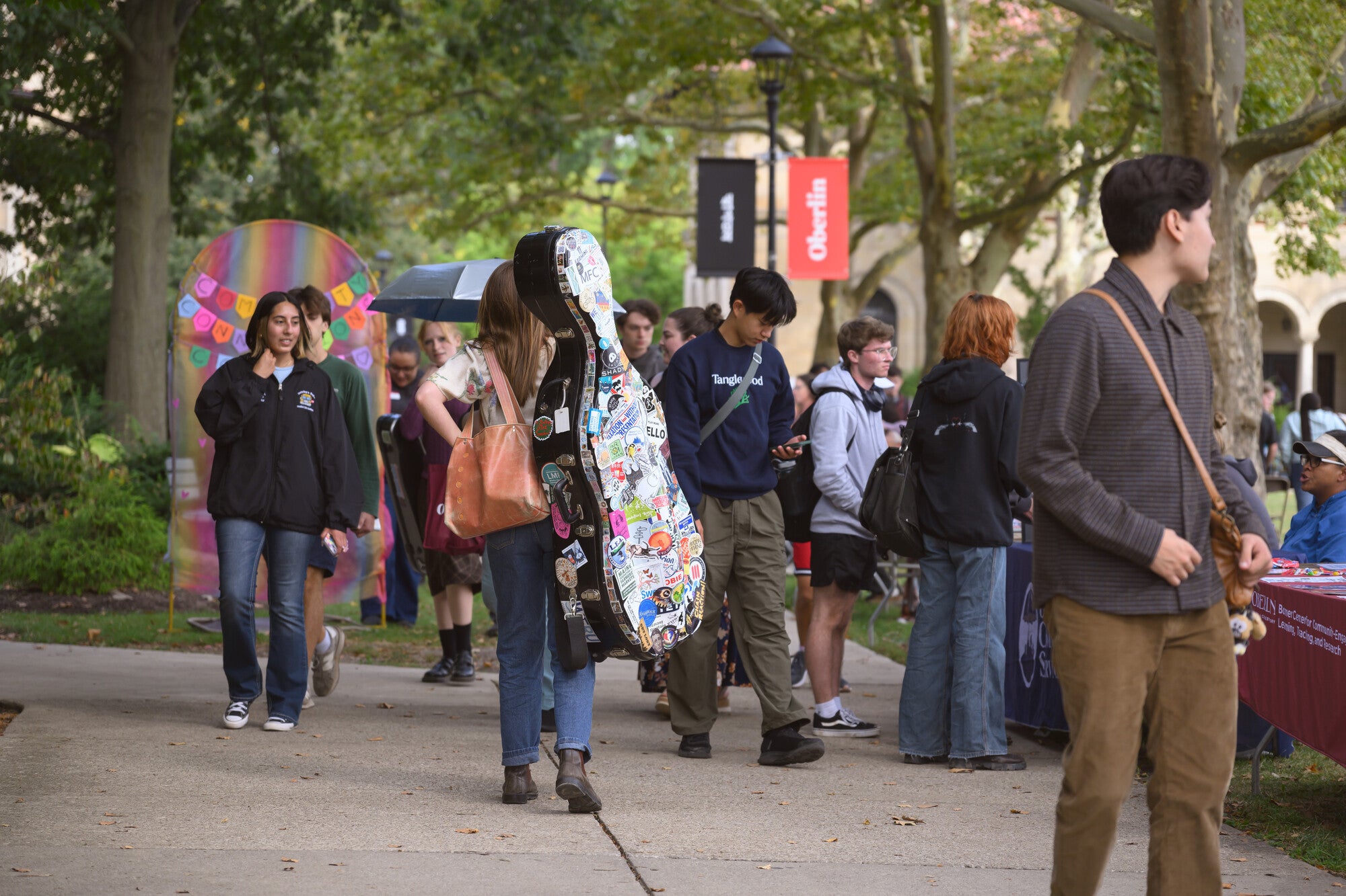 People walking along a tree-lined campus path, some carrying musical instruments.