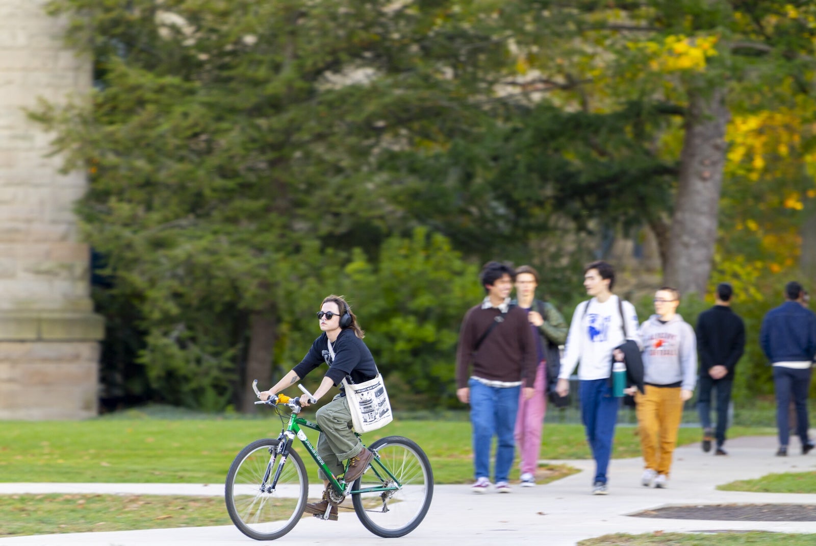 Student riding a bicycle on campus with other students walking nearby.