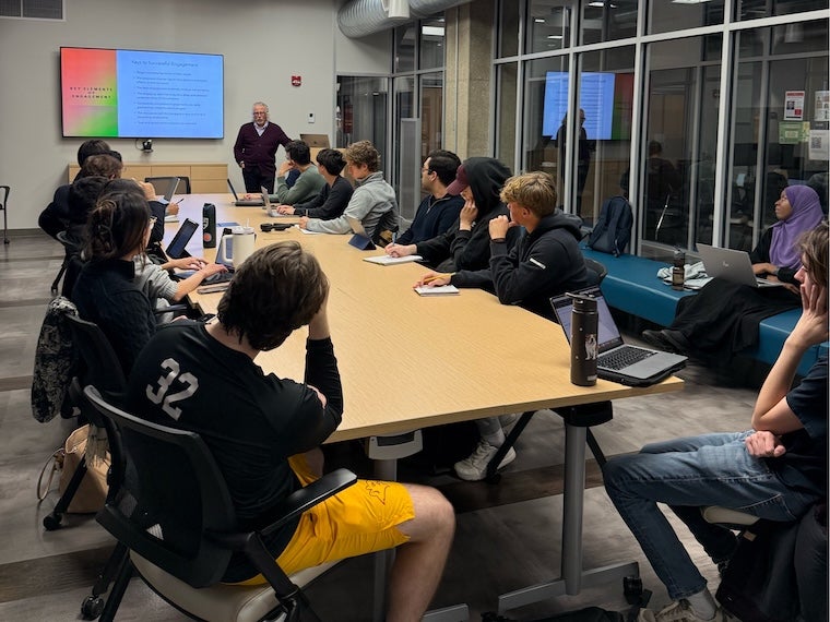 Students and faculty seated around a conference table in a classroom meeting, with presentation screens visible in the background.