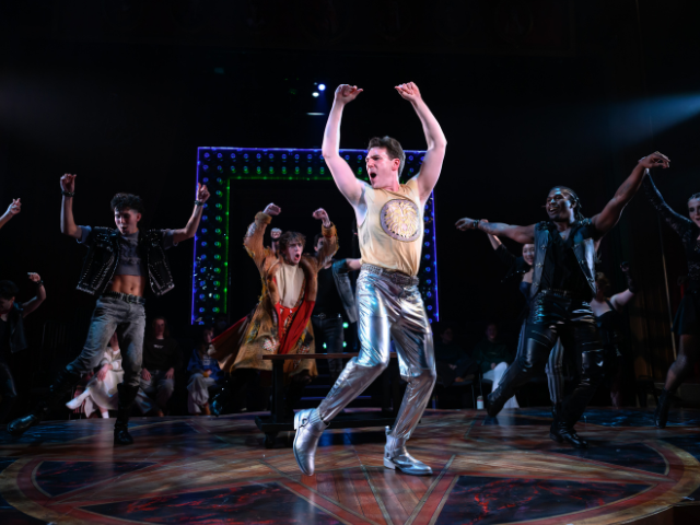 Stage performance with a central dancer in a gold top and silver pants raising both arms, surrounded by other performers dancing under colorful stage lights.