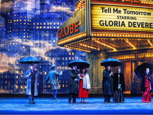 **Alt text:** Stage scene of a rainy city street outside the Globe Theatre, where several people stand with umbrellas beneath a glowing marquee reading “Tell Me Tomorrow Starring Gloria Devere,” including a woman in a red dress at center.