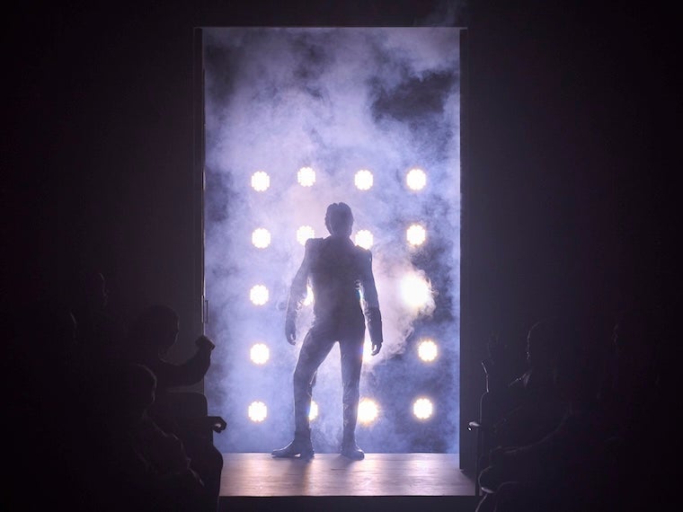 Silhouetted performer standing in a doorway framed by bright stage lights and theatrical haze.