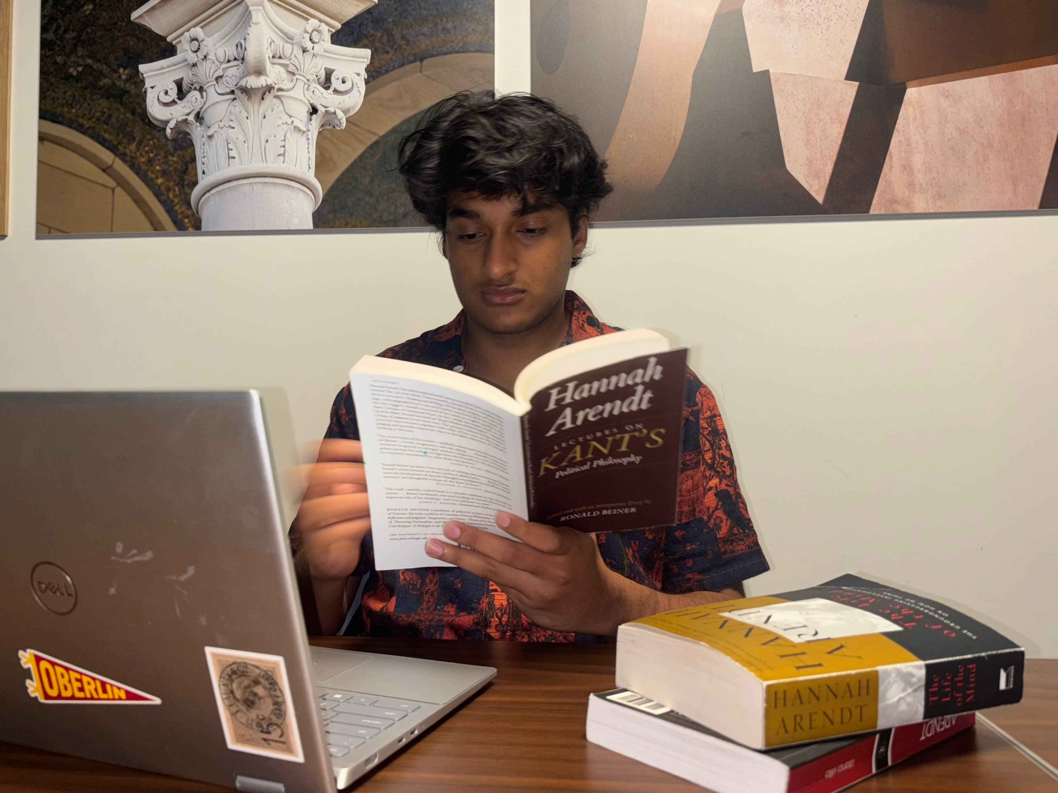 Harsh reads Hannah Arendt's book "Lectures on Kant's Political Philosophy" while seated at a desk behind a laptop and stack of two books.