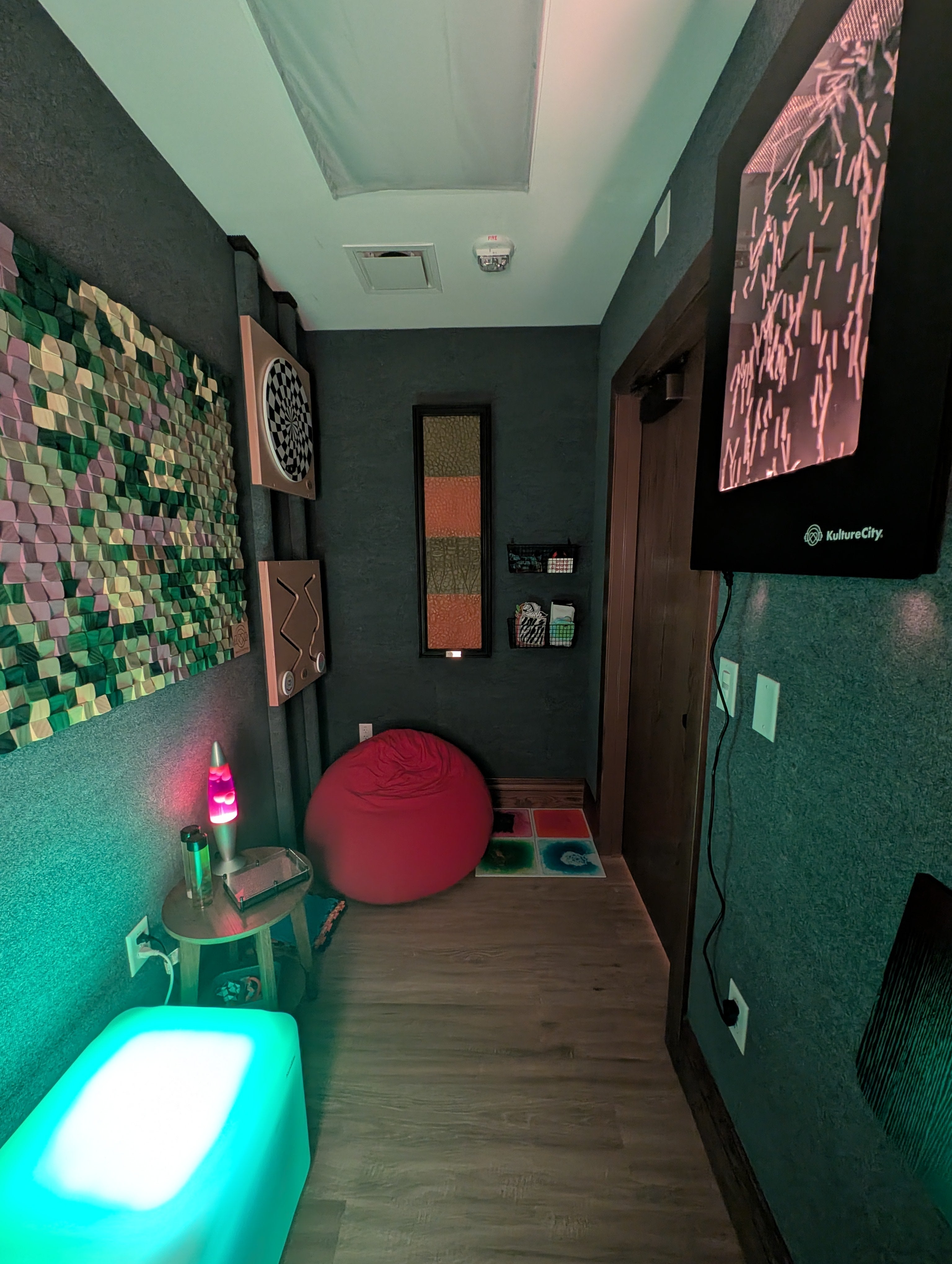 The sensory room is dimly lit by a glowing square block, showing a soft beanbag chair and multiple sensory boards on the walls.
