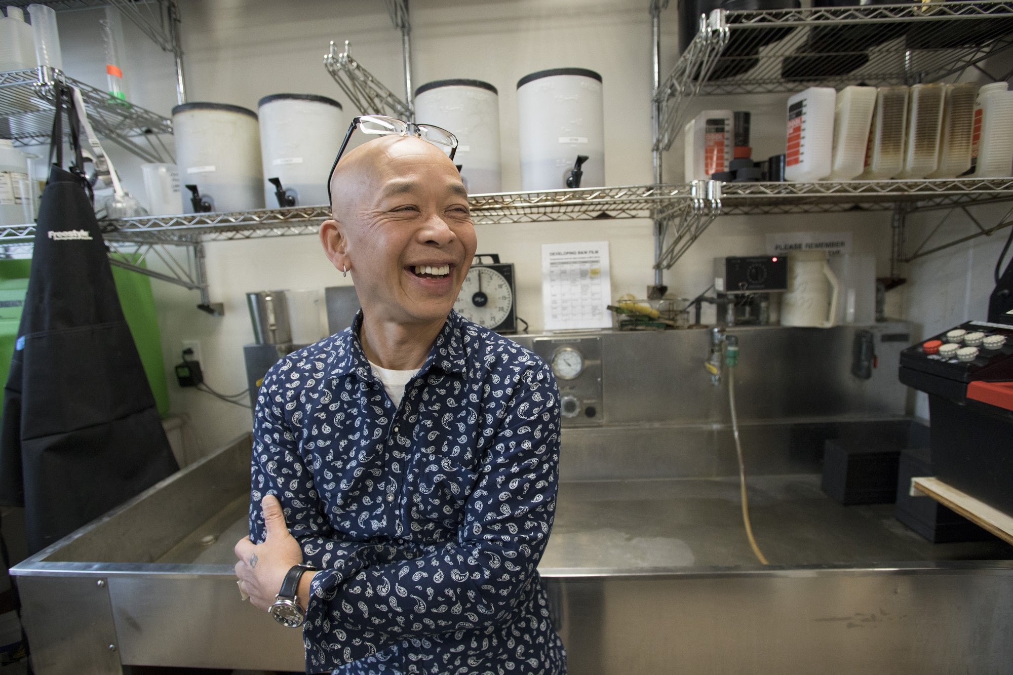 Pipo Nguyen-duy, in a blue paisley shirt with glasses pushed up on his head, laughing with his arms crossed in front of a steel sink in the Oberlin photography darkroom.