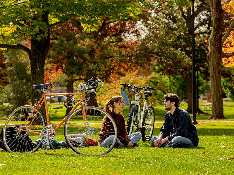 Two students sit in the grass talking, with their bikes standing nearby.