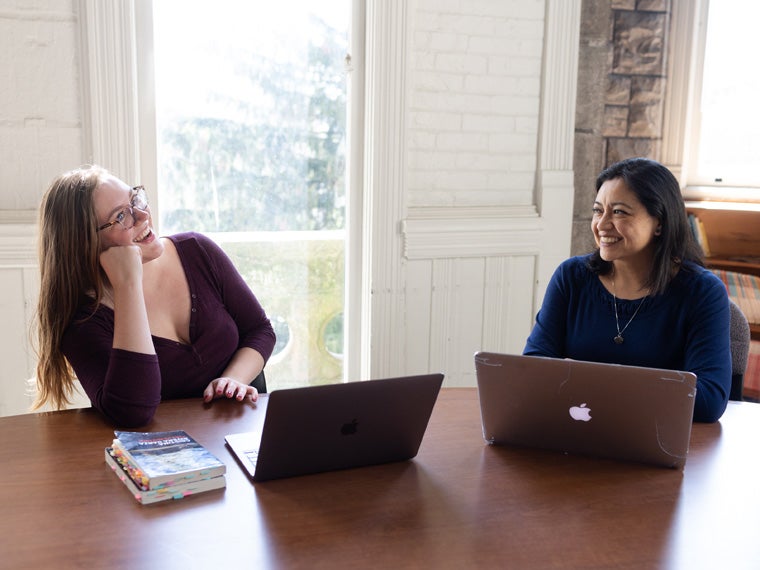 Two women at a table with open computers, sharing a laugh.