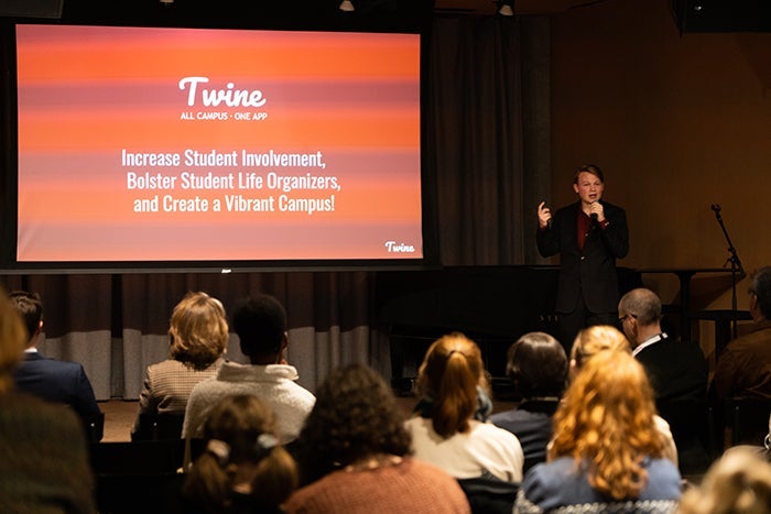 A student presenter at the podium. The screen shows a logo, Twine.