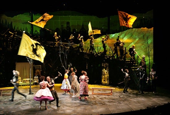 A stage full of actors. On the floor, women in dresses are dancing with men in suits. Above on a staircase, soldiers are holding flags with a bird.