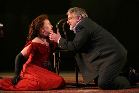 Two actors in an emotional interaction. The woman, wearing a red dress, is sitting on the ground gazing at a man who is kneeling next to her with his hand on her chin