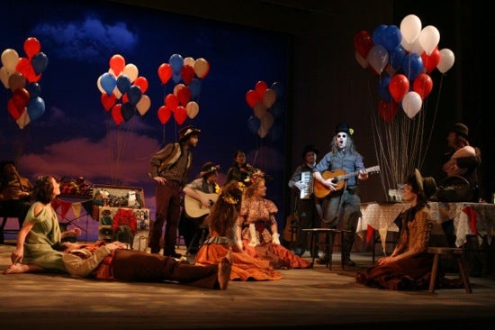 A stage full of actors. They are listening to a man sing and play guitar. They are surrounded by red, white, and blue balloons