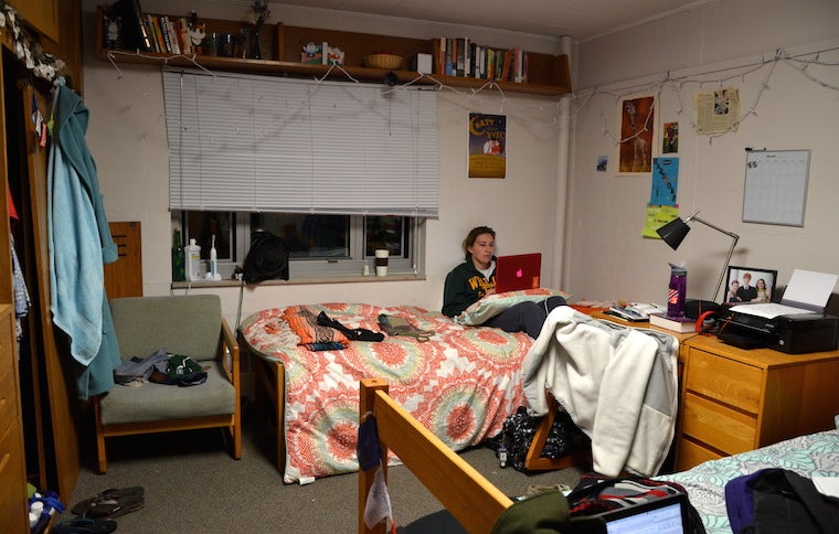 A girl sitting on a bed in an open-double dorm room. The girl has her feet propped up on her desk and is looking at her lap-top. 