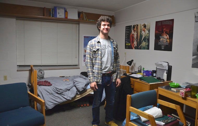 A boy standing in a dorm room looking at the camera. 