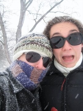 two people posing for a selfie. Both are wearing sunglasses and gear for cold weather. It is snowing behind them and they are covered in snow flakes. 