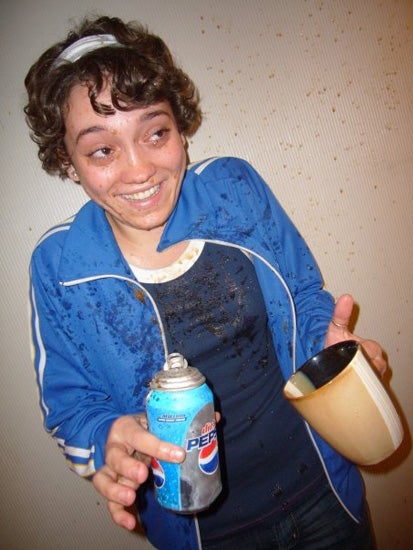 A girl holding an open Pepsi can and a coffee mug. She is covered in what looks to be sprayed Pepsi