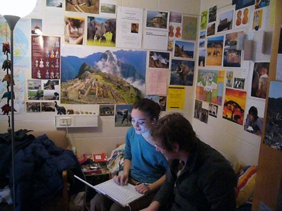 Two people sitting on the bed of a dorm room looking at a laptop. The wall behind them is full of posters