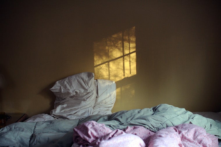 An unmade bed, the reflection of sun coming through a window onto a wall 