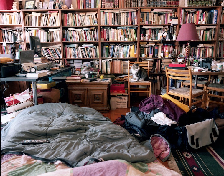 A room full of bookshelves, desks, a cat, a suitcase, and a make-shift bed on the floor