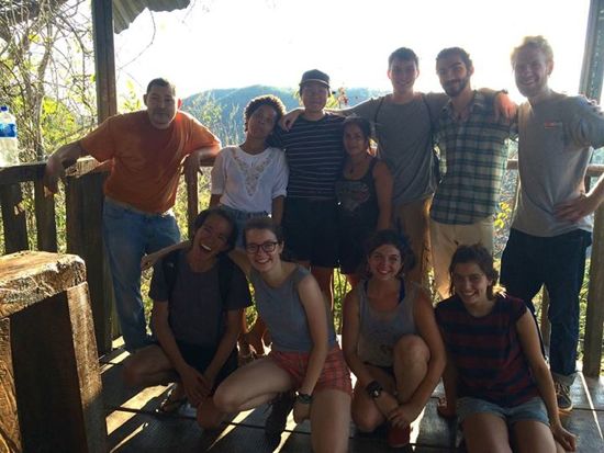 A group of students pose with their arms around each other at a railing overlooking mountains 