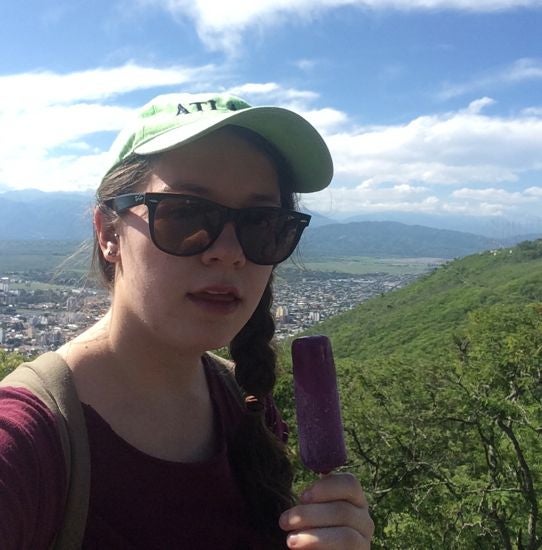 On a mountain with a town behind her, Esther eats a grape popsicle.
