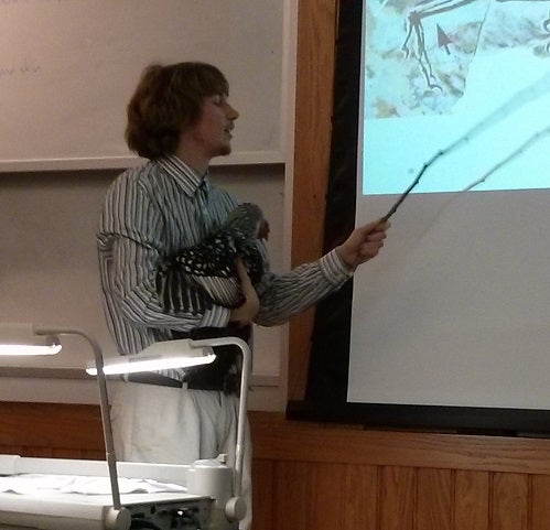 A professor speaks to a powerpoint while holding a chicken 