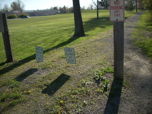 A chain connected by two short wooden pillars blocking a dirt path. A sign hangs from the chain: "park closes at dusk." Another sign: "No parking anytime"