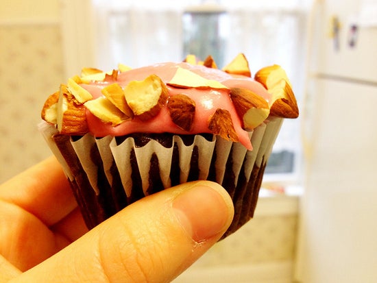 Fingers holding a tiny brown cupcake with pink frosting and almond bits