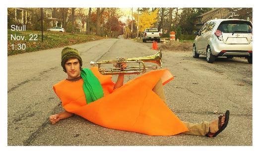 student wearing a carrot costume laying in the road holding a brass instrument 