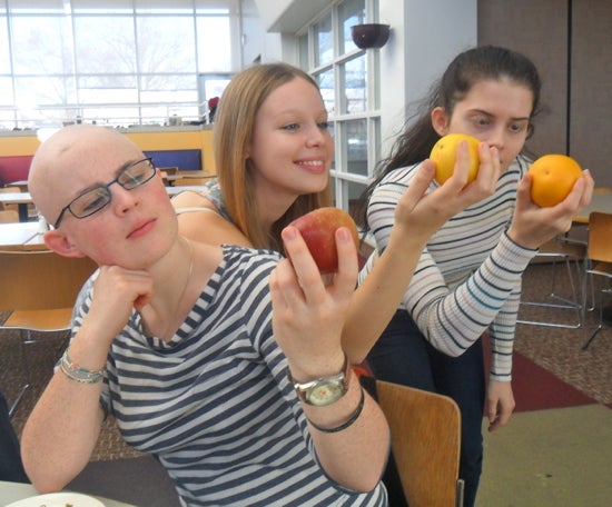 Three students hold apples and oranges as they gaze at the fruit