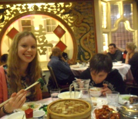 Author poses for the photo holding food with chopsticks and her brother is mid bite 