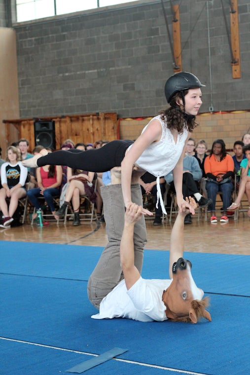 Lying on his back on a mat and wearing a horse head mask, Teague holds a girl aloft on his raised feet.