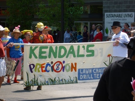 A sign announces the next group: Kendal's Precision Lawn Chair Drill Team.