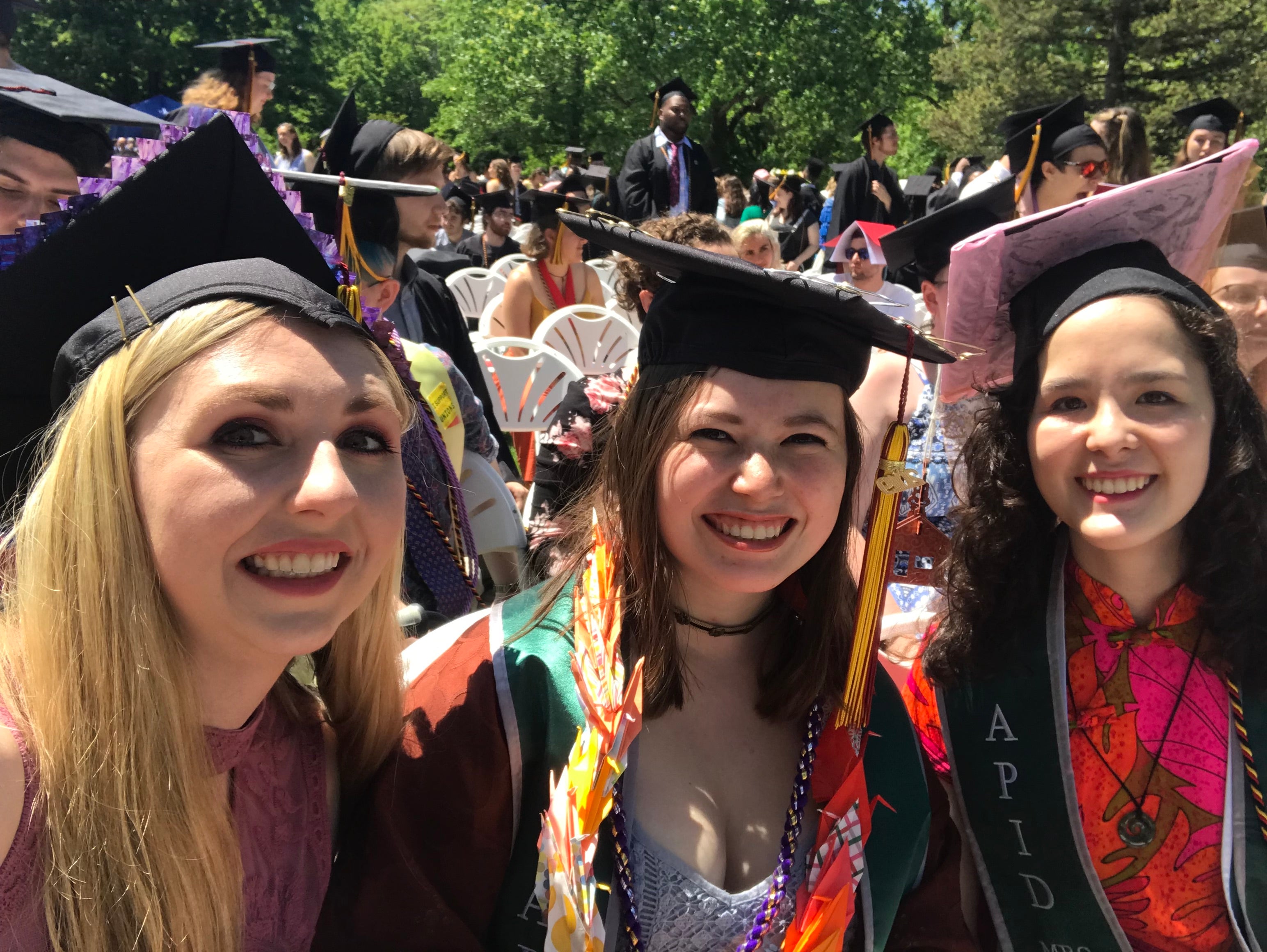Three women wearing graduation caps. One is blond with purple on her cap, the other is brunette and wears a necklace made of paper cranes, and the other has curly brown hair and is wearing a pink cap and dress.