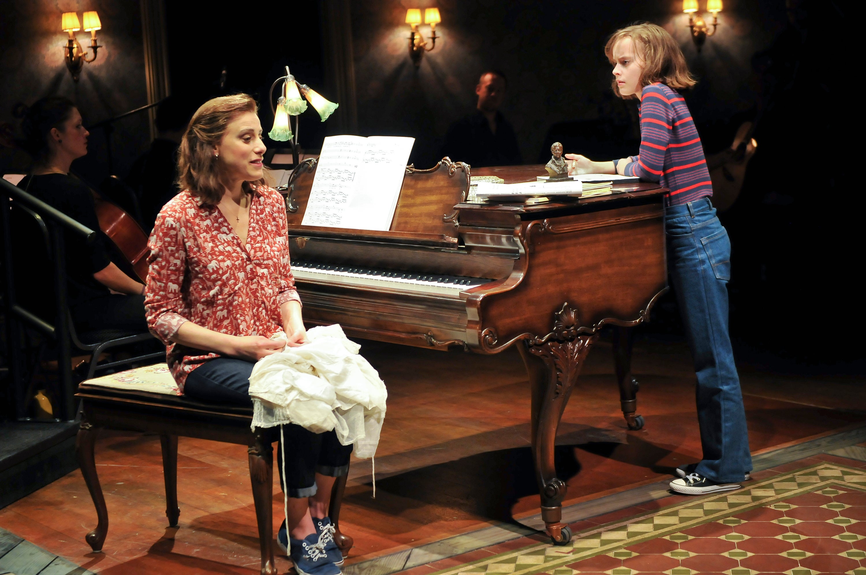 Judy Kuhn sitting next to a piano in onstage in Fun Home. Sydney Lucas leans on the piano