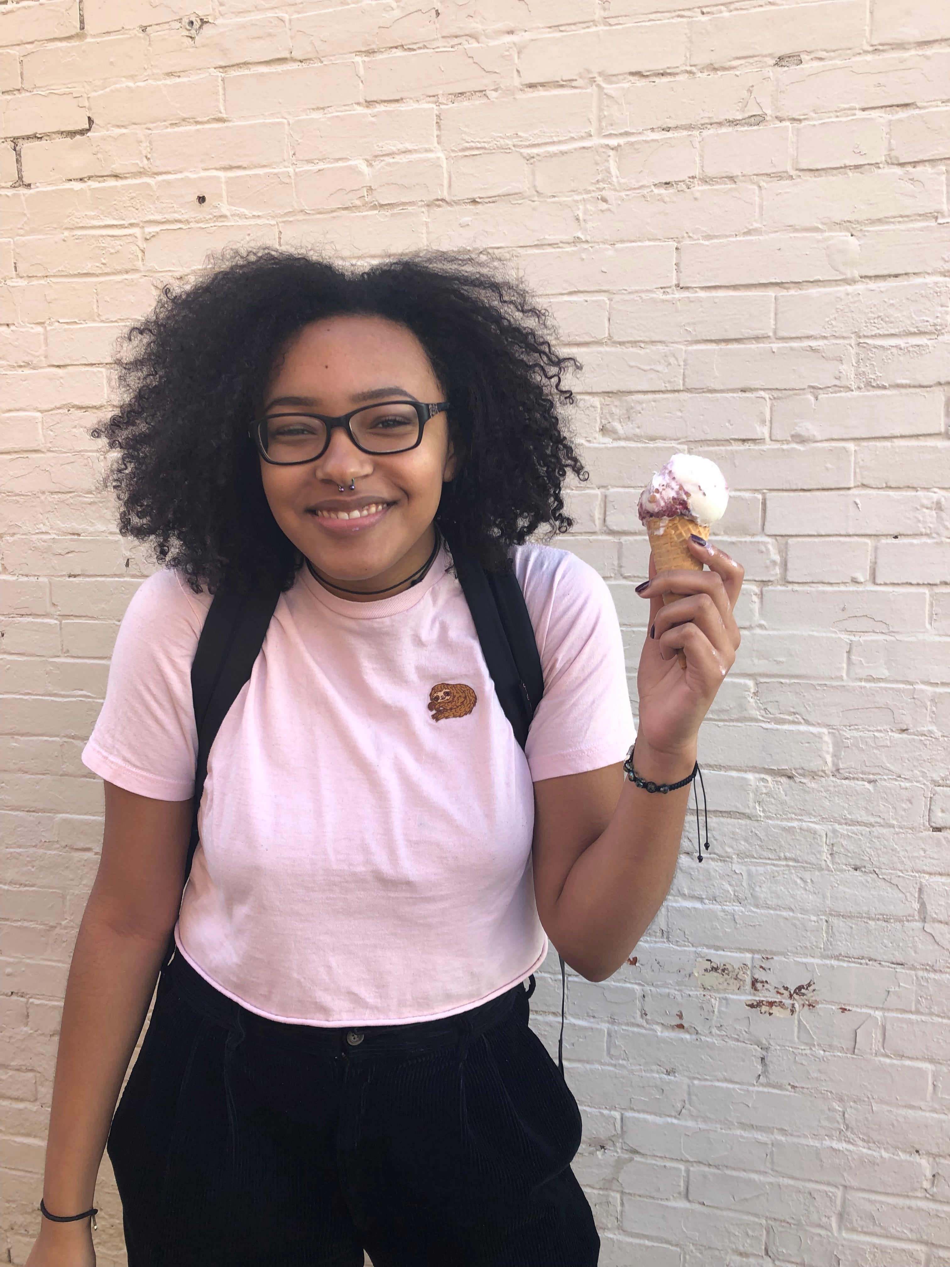 Imani smiling with an ice cream in her hand, a pink crop top, and black pants, standing against a light yellow brick wall.