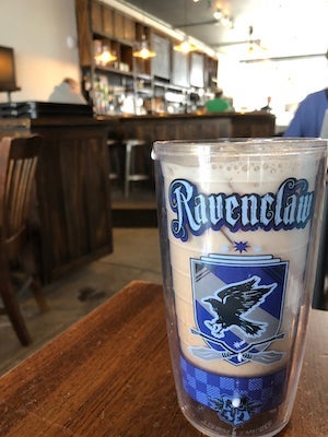 A milky chai latte with small cubes of ice sits in a blue reusable cup with 'Ravenclaw' written across it in blue letters, with the Harry Potter crested below it. Behind the drink are hanging lights and a dark brown, rustic-looking counter and silver stools.