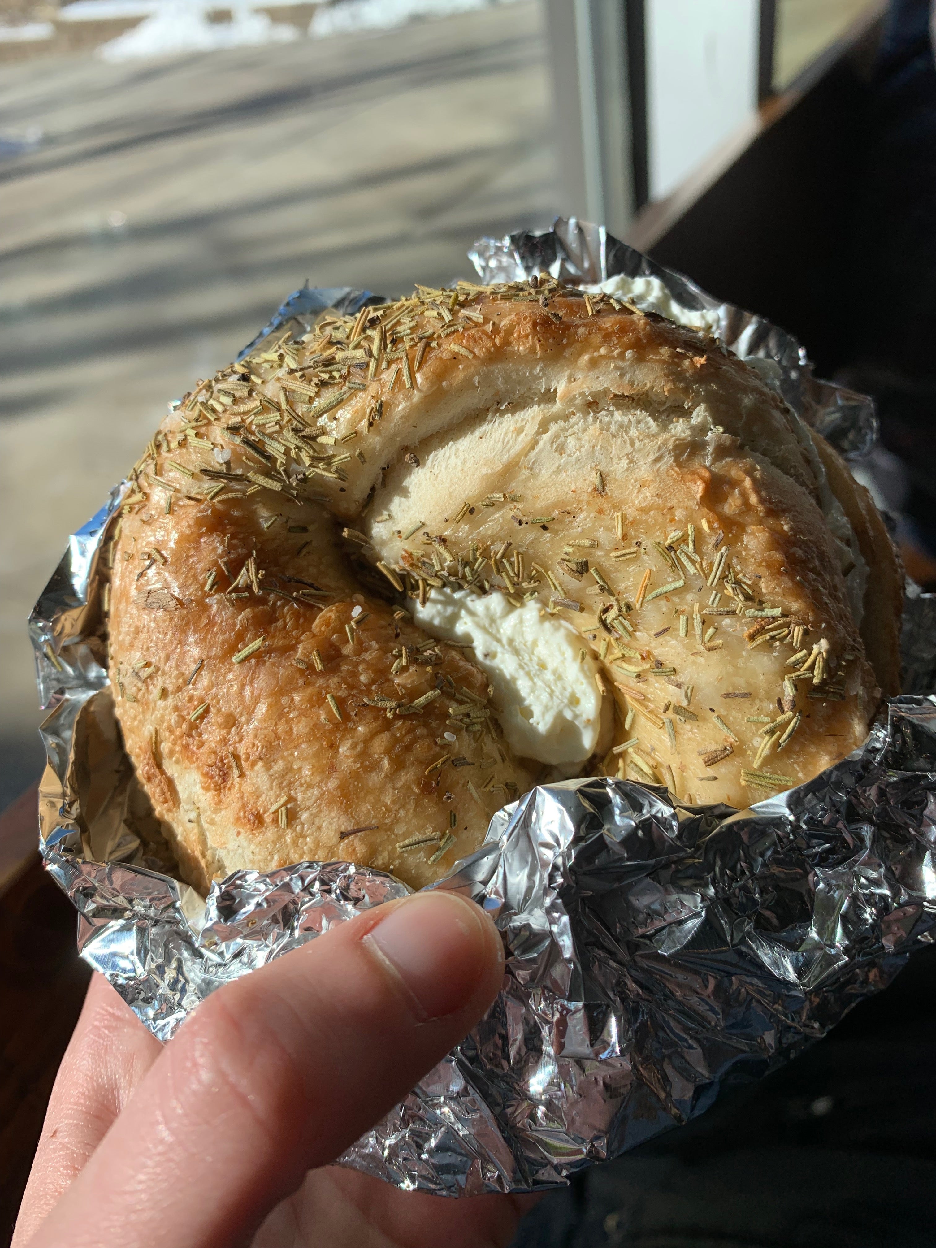 a hand gently caresses a bagel with rosemary and cream cheese, wrapped in foil, on a sunny day